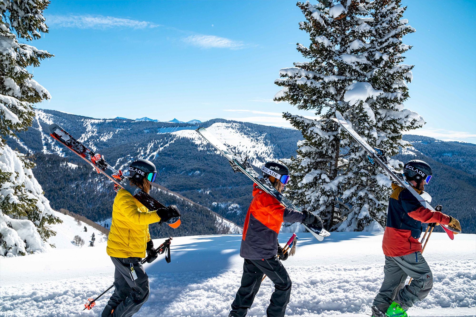 Three people in winter gear carry skis on a snowy mountain with a scenic view of trees and hills in the background.