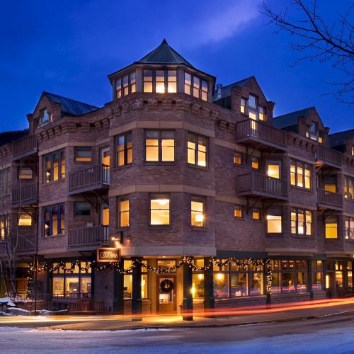 A warmly lit, multi-story building at dusk with a turreted roof. Light trails from passing cars are visible in the foreground.