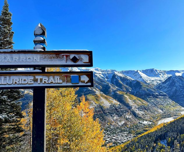 Mountain landscape with a wooden signpost pointing to "Cimarron Ridge" and "Telluride Trail" amidst fall foliage and snow-capped peaks.