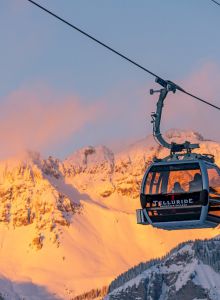 A gondola labeled "Telluride" moves across snowy mountains at sunset, with pinkish skies creating a picturesque winter scene.