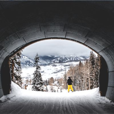 A skier in yellow pants stands at the tunnel's exit, overlooking a snowy landscape with trees and mountains in the background.