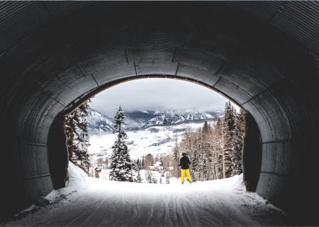 A skier in yellow pants stands at the tunnel's exit, overlooking a snowy landscape with trees and mountains in the background.