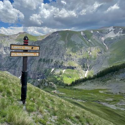 Trail signs on a mountain path with a scenic view of green slopes and clouds in Telluride Colorado .