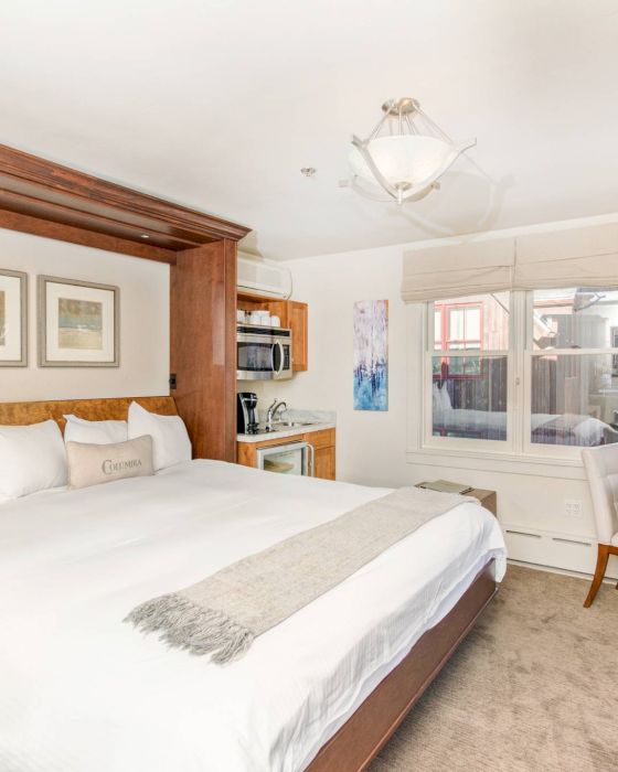 A well-lit Hotel Columbia Telluride bedroom with a large bed, bedside tables, lamps, artwork, and a window.