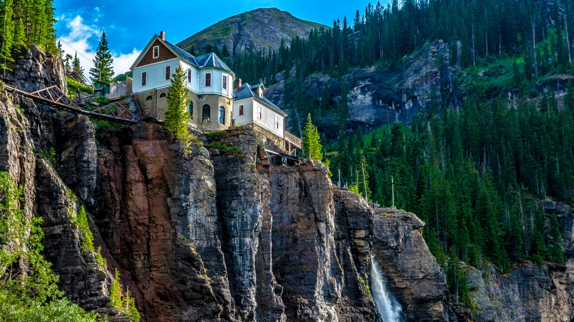 A house perched atop a cliff with a waterfall flowing nearby, surrounded by lush greenery.