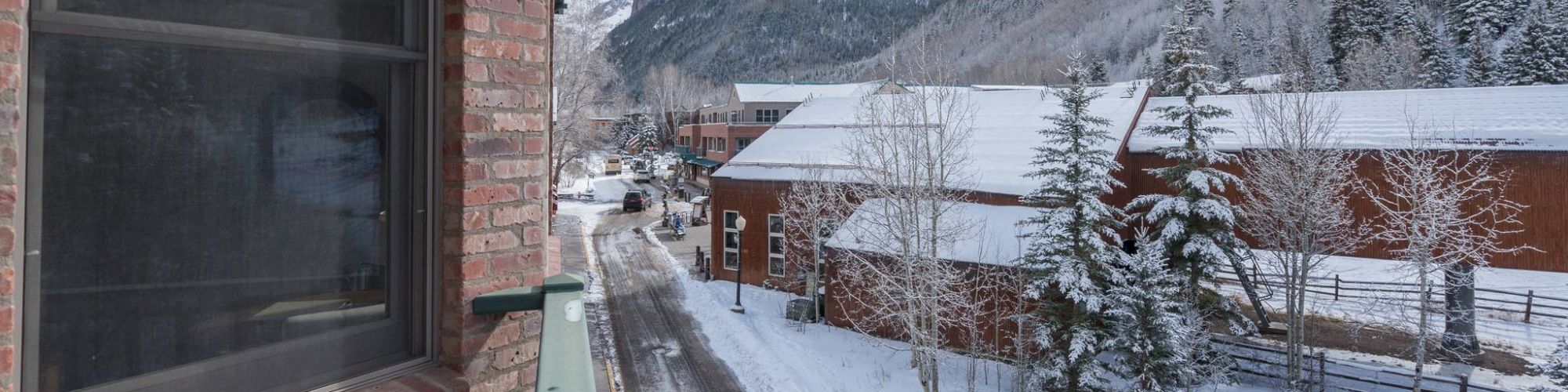 Snow-covered street with buildings, mountains in the background, and a person walking.