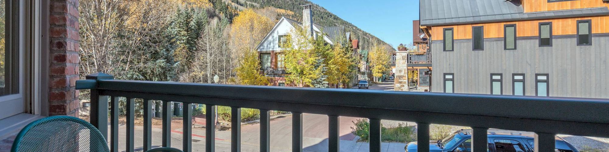 View from a balcony featuring chairs, overlooking a street, buildings, and autumn trees against a mountain backdrop.