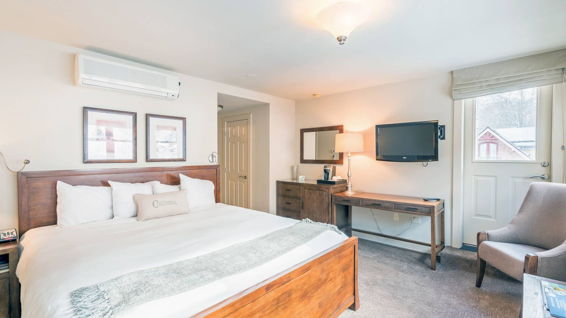 A neat Hotel Columbia Telluride bedroom with a bed, armchair, desk, TV, and bright lighting.