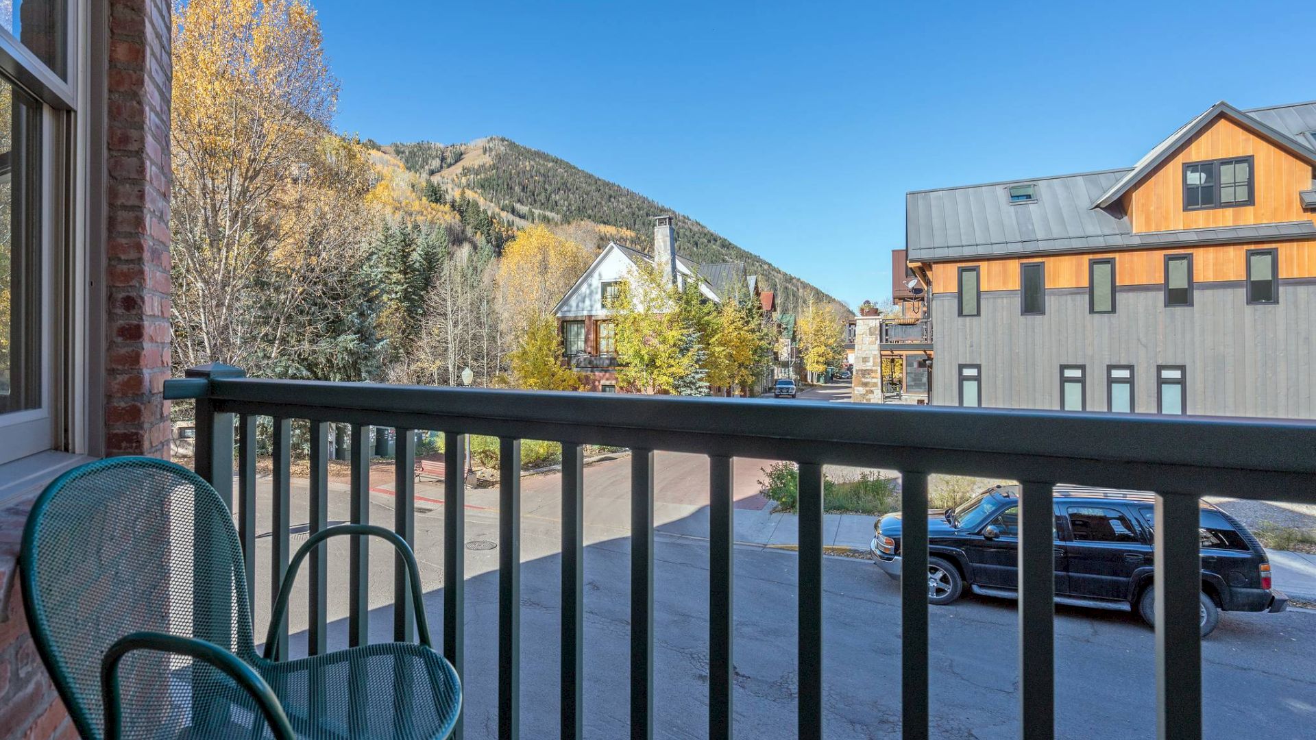 Balcony view of a mountain town with autumn trees, buildings, a chair, and parked cars.