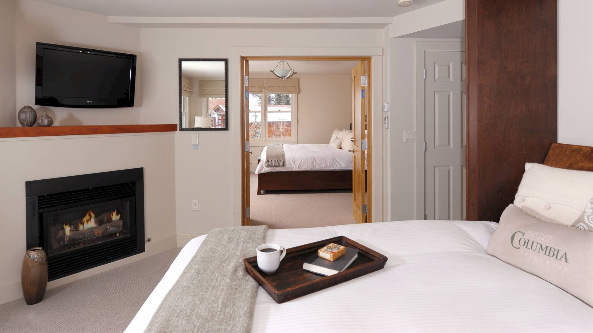 Cozy bedroom with a fireplace, TV, and a tray with food on the bed, connecting to another room.