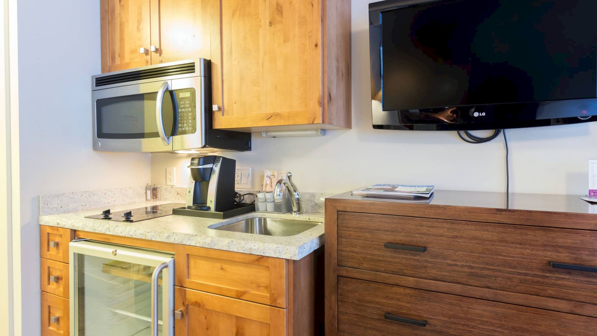 A kitchenette with wood cabinets, microwave, sink, fridge, and a mounted TV.