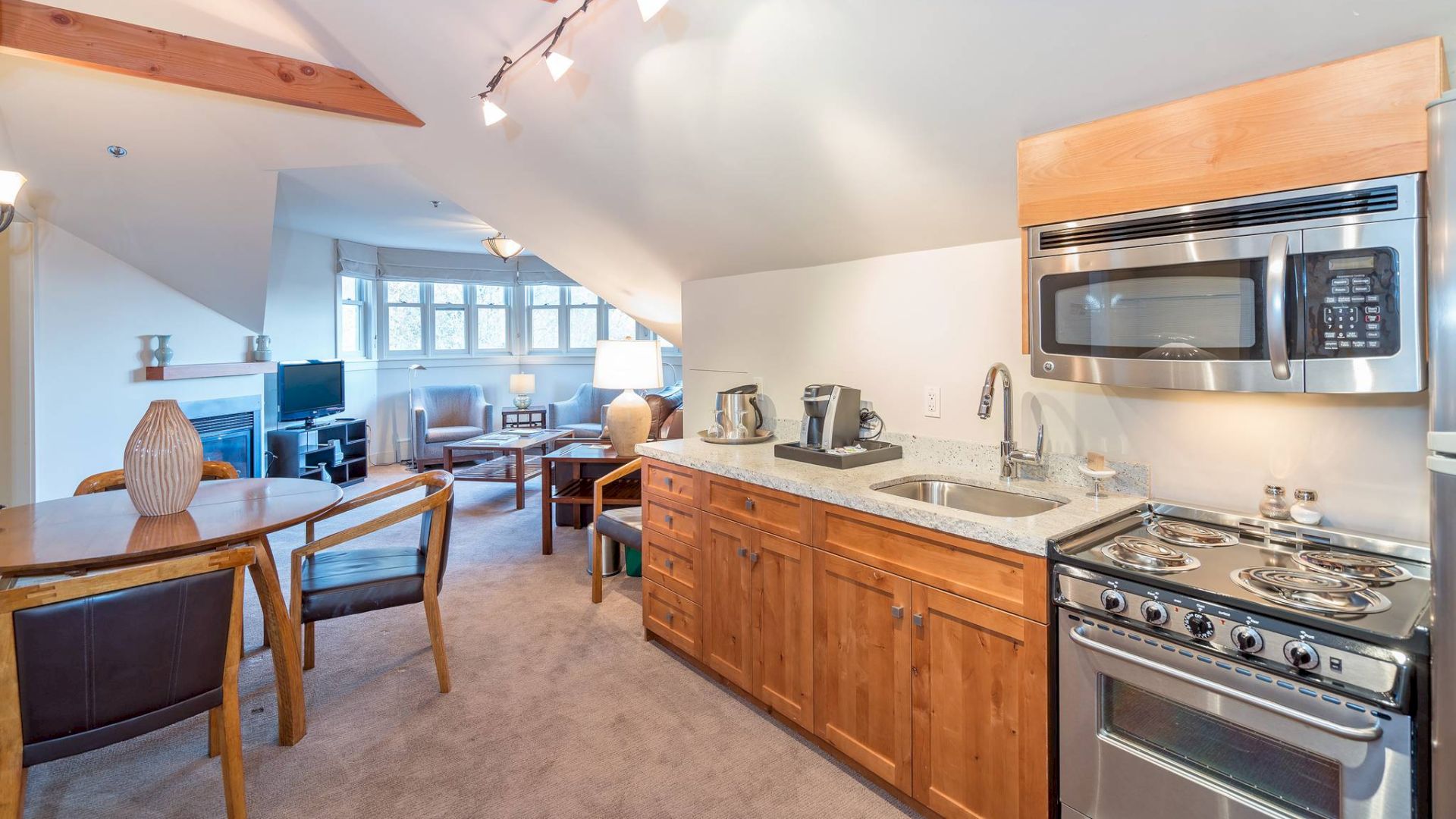 A modern kitchen with stainless appliances, wooden cabinets, and a dining area leading to a living room space.