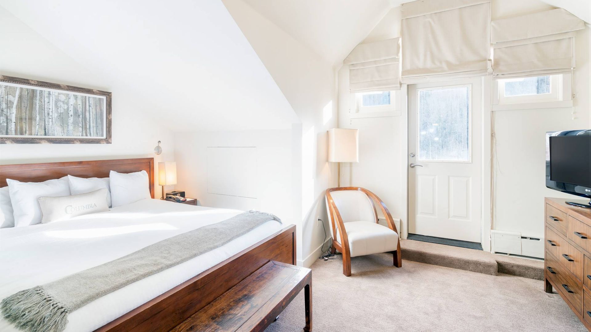 Cozy bedroom with a bed, dresser, lamp, and a chair under sloped ceiling.