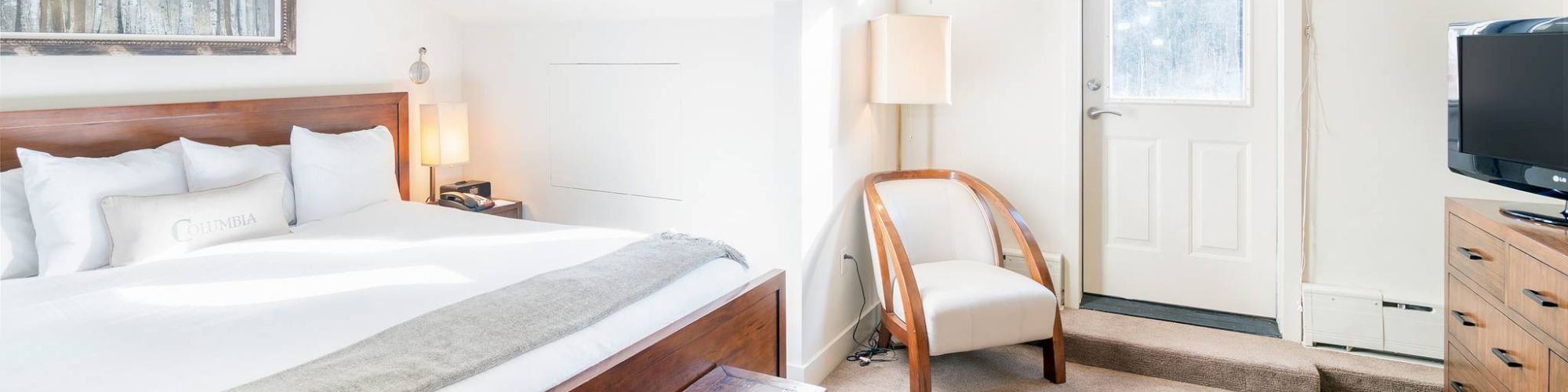 Cozy bedroom with a bed, dresser, lamp, and a chair under sloped ceiling.