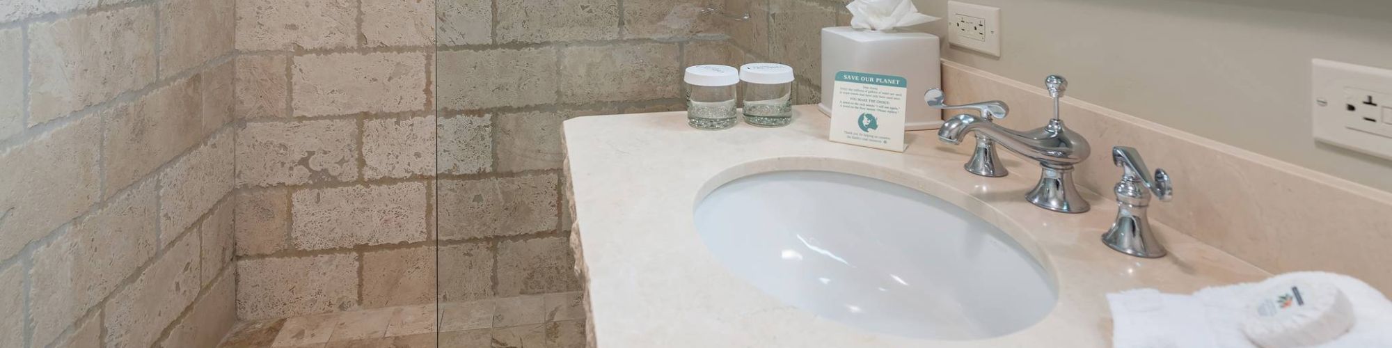 A bathroom sink with toiletries and towels, surrounded by beige tiling.