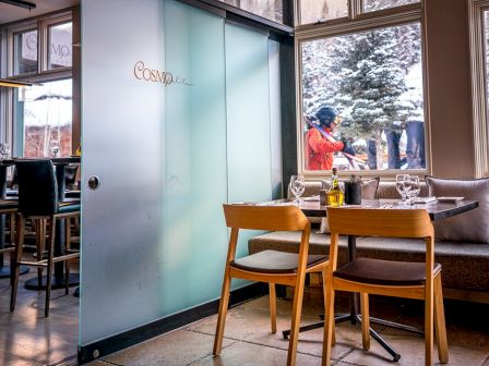 Inside a restaurant with tables, chairs, a set table, and a person outside by the window.