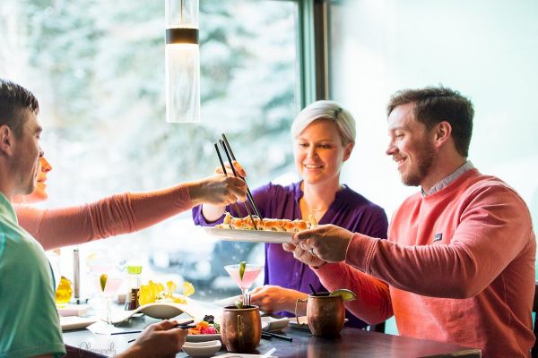 Three people are enjoying a meal together, sharing food, in a bright setting.