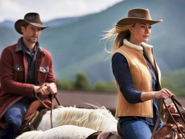 Two people on horseback, wearing hats and outdoor clothing, with a mountainous backdrop.