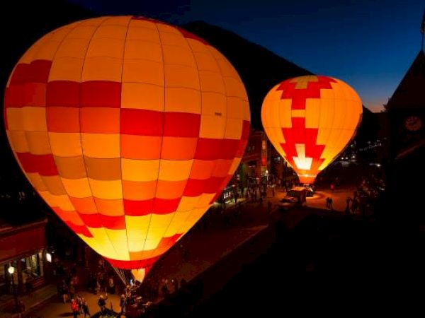 Illuminated hot air balloons glow at dusk with a crowd and buildings nearby.