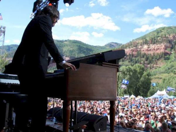 A musician plays a piano at an outdoor festival with a crowd and mountains behind.