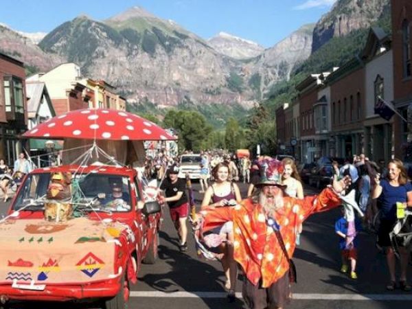 A colorful street parade with a festively decorated car and people in costumes, against a mountain backdrop.