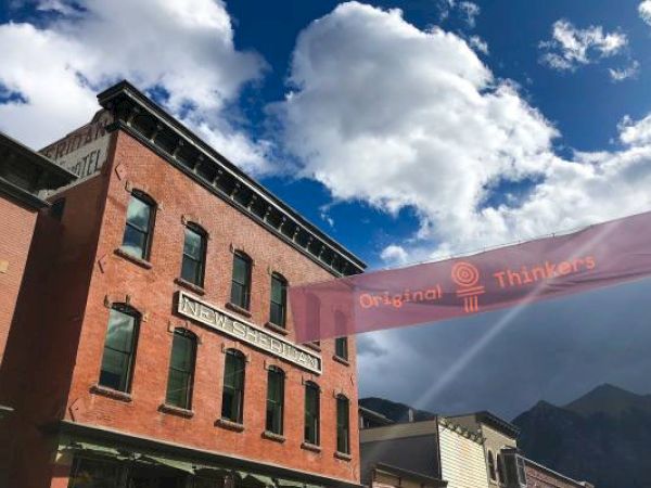 Brick building under a blue sky with clouds, a banner reading "Original Thinkers," and mountains in the background.