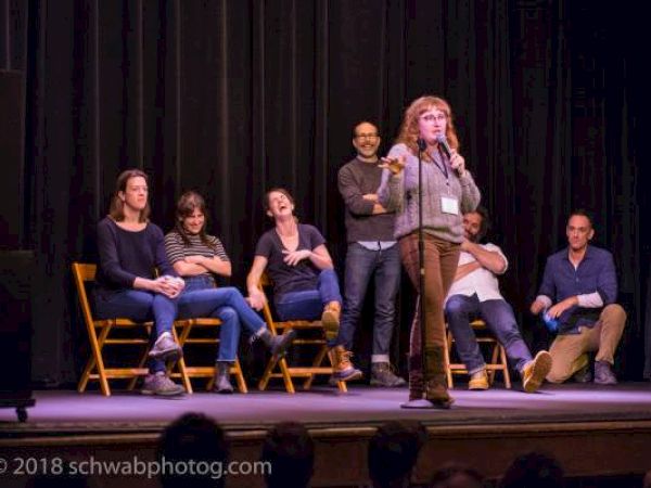 A person speaks into a microphone on stage, with others seated behind laughing.