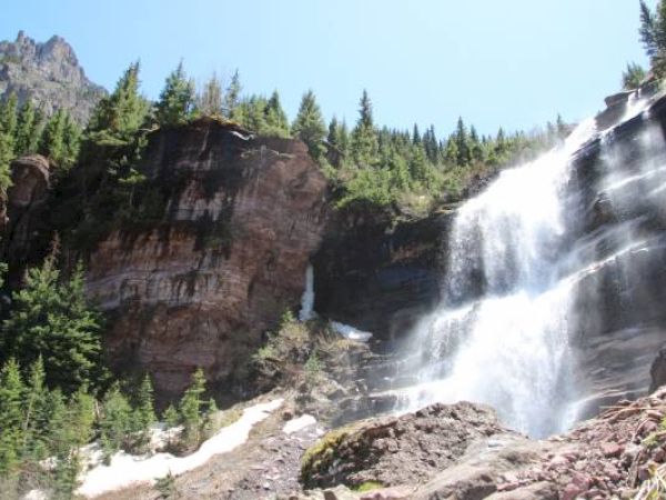 A picturesque waterfall cascades down a rocky cliff surrounded by lush greenery and clear skies.