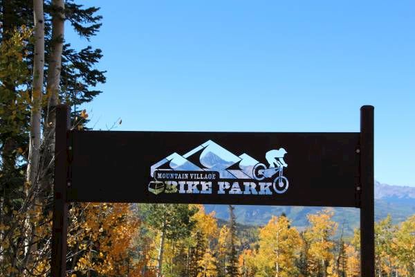 A mountain village sign with a cyclist, trees in autumn colors, clear blue sky.