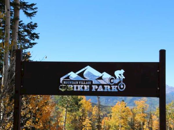 A mountain village sign with a cyclist, trees in autumn colors, clear blue sky.
