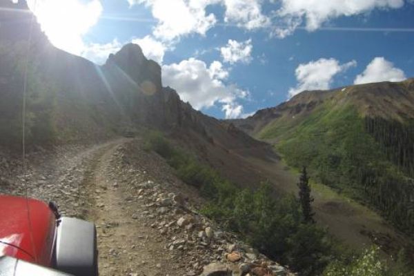 Rugged mountain road with a red vehicle's hood, surrounded by greenery under a sunny sky.