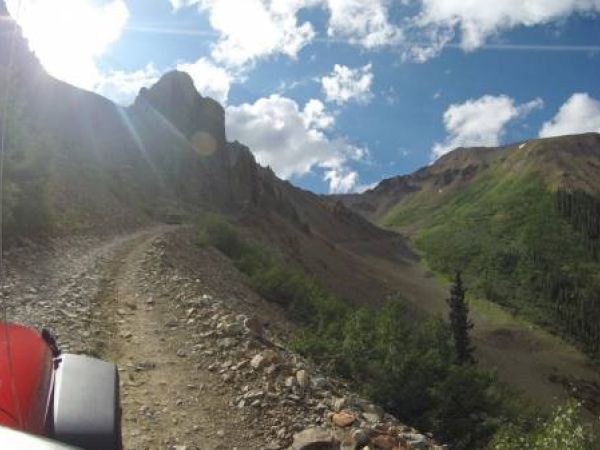 Rugged mountain road with a red vehicle's hood, surrounded by greenery under a sunny sky.