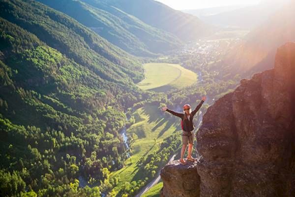 A person stands on a cliff edge with arms outstretched, overlooking a verdant valley bathed in sunlight.