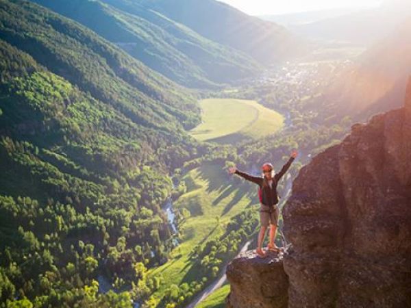 A person stands on a cliff edge with arms outstretched, overlooking a verdant valley bathed in sunlight.