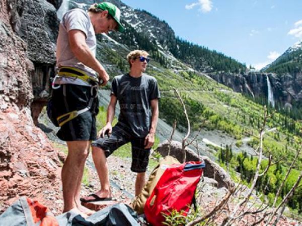Two climbers with gear stand by a cliff with a scenic mountain backdrop and waterfall.