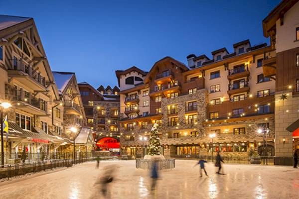 Twilight at a cozy ice rink in a festive village square with buildings around.