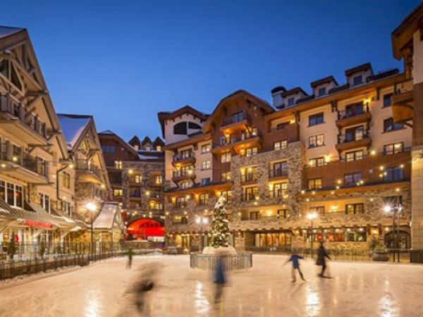 Twilight at a cozy ice rink in a festive village square with buildings around.