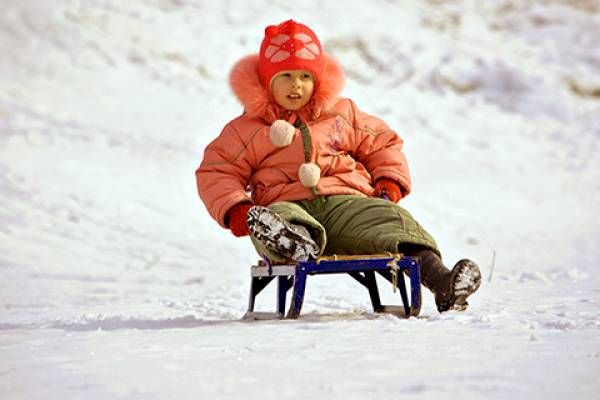 A child in warm winter clothes sits on a sled in the snow.