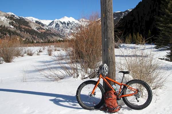 An orange fat tire bike leans on a pole in a snow-covered landscape with mountains in the background.