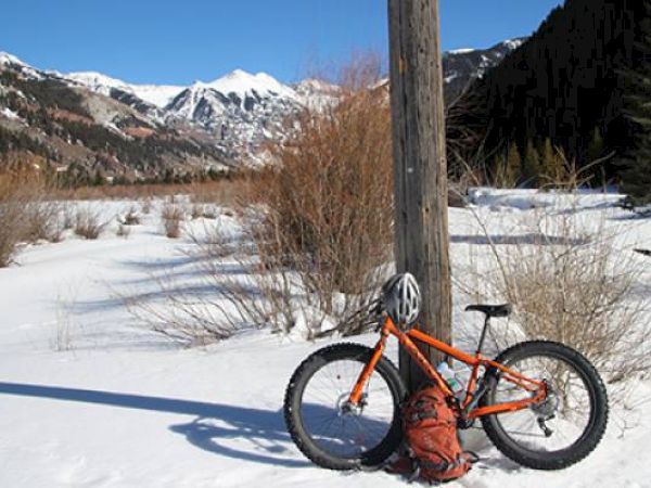 An orange fat tire bike leans on a pole in a snow-covered landscape with mountains in the background.