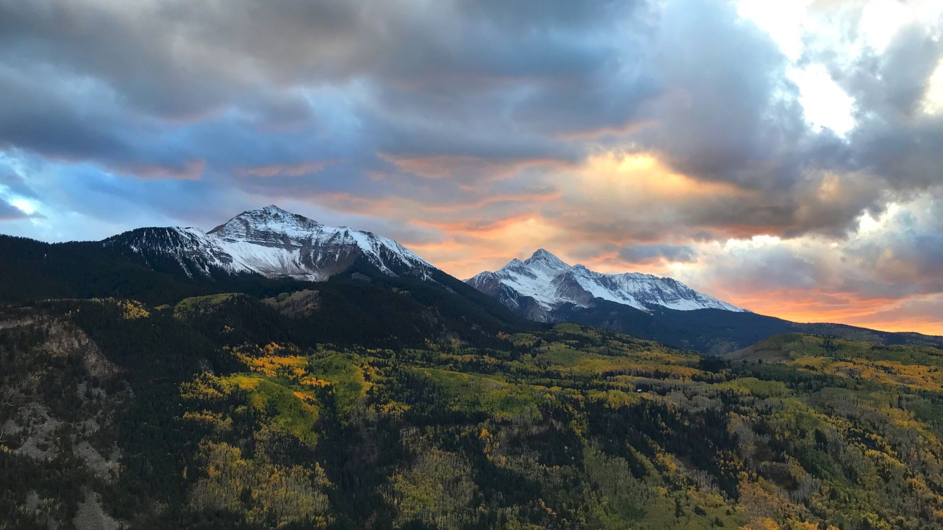 Mountain peaks with snow under dramatic sunset skies, foreground of autumn foliage.