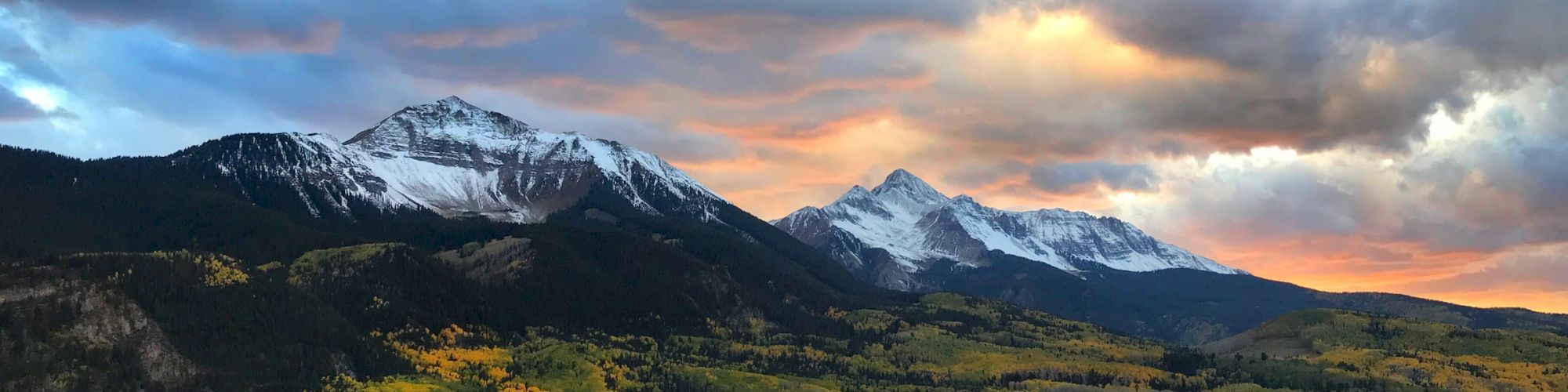 Mountain peaks with snow under dramatic sunset skies, foreground of autumn foliage.