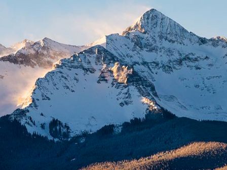 Snow-covered mountains with sunlight touching the peaks, and forested hills at sunrise or sunset.