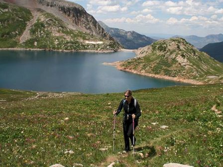 A hiker with poles near an alpine lake and hills under a blue sky.