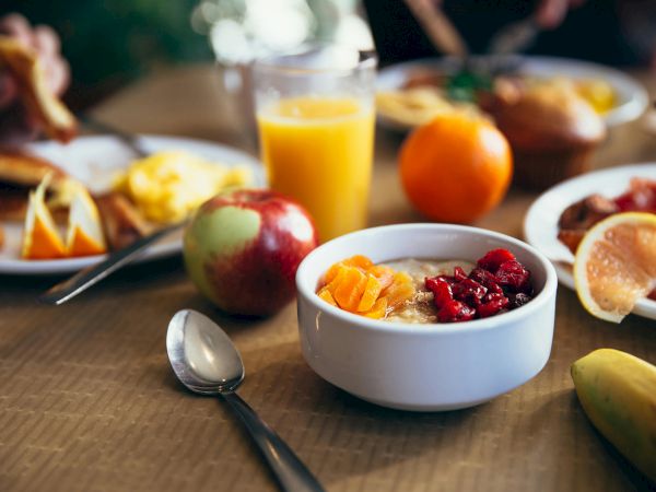 A breakfast scene with oatmeal, fruit toppings, apple, orange, juice, scrambled eggs, and toast on plates, and a spoon on the table.