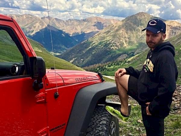 A man leans on a red Jeep with mountains in the background.