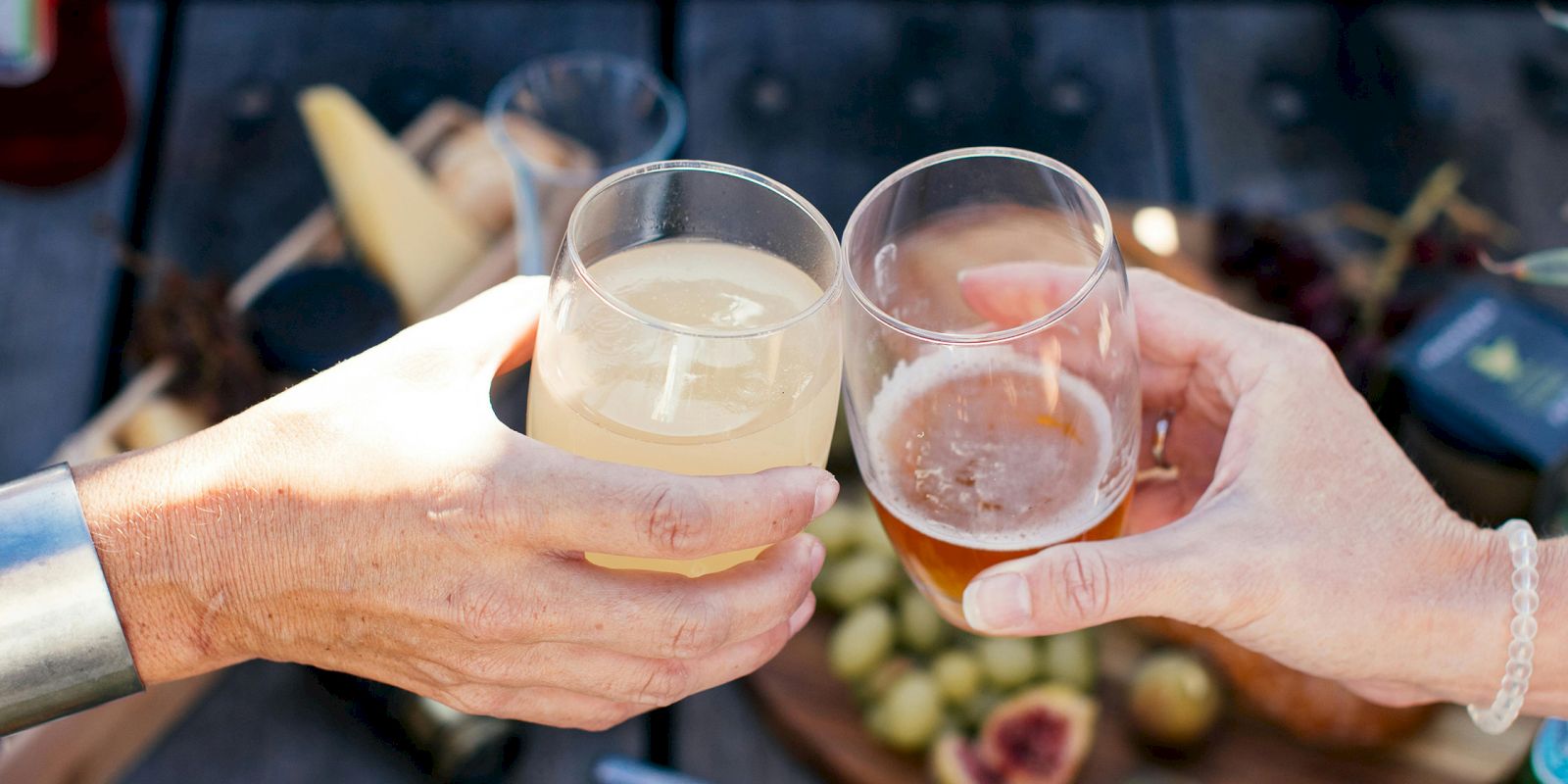 Two hands clinking glasses above a picnic spread with cheese and fruit.