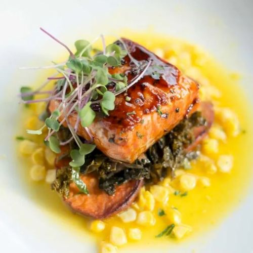 Grilled salmon fillet on top of greens with corn and a yellow sauce, garnished with microgreens.