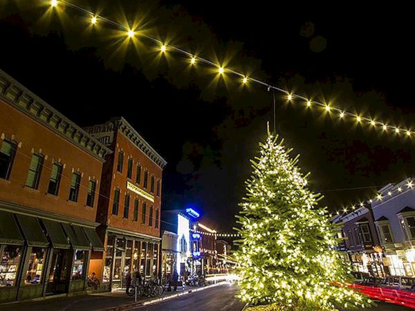 A night view of a festive street with buildings, string lights, and a large illuminated Christmas tree.