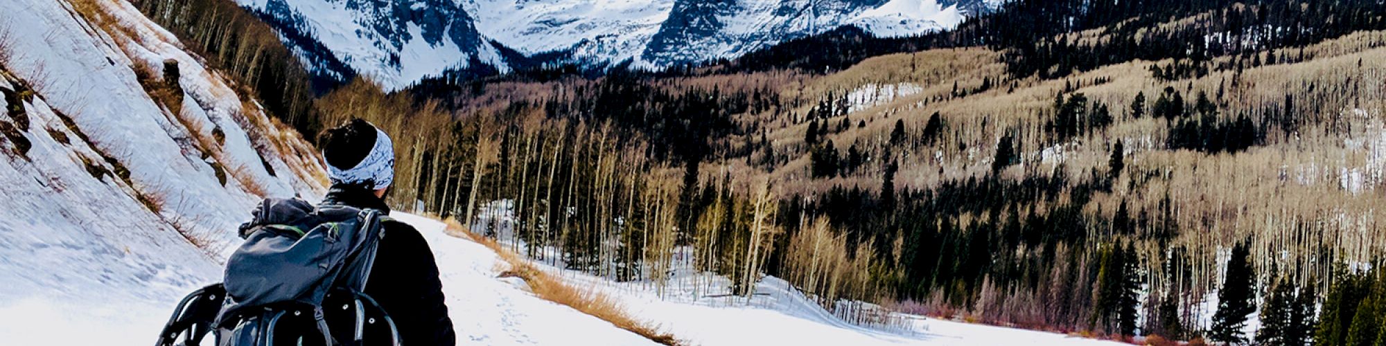 A person with a backpack walks through a snowy landscape, surrounded by mountains and trees under a partly cloudy sky.